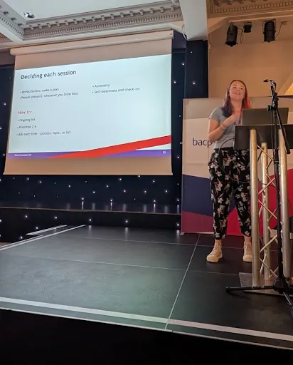 Becca standing on stage behind a podium; giving a talk. A presentation is projected
off to the left