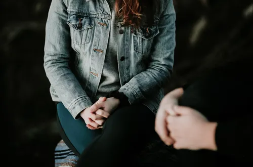 A person sat crossed legged on a stool facing the camera, with another person facing them off to the side